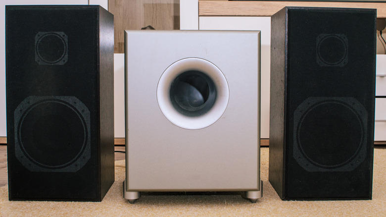 A silver, modern looking subwoofer sits between two black speaker cabinets in what appears to be the inside of a white room