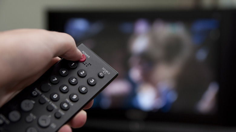 Hand pressing the power button on a TV remote with a blurred television in the background.