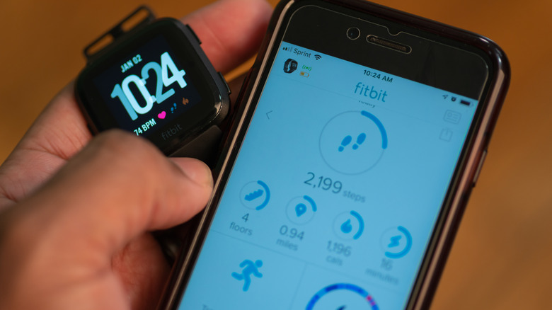 A hand holding a Fitbit smartwatch and a phone showing the app.