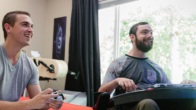 Two men sitting next to each other playing video games. The one on the right has a white Xbox controller, while the one on the right has a customized set-up using the Xbox Adaptive Controller.