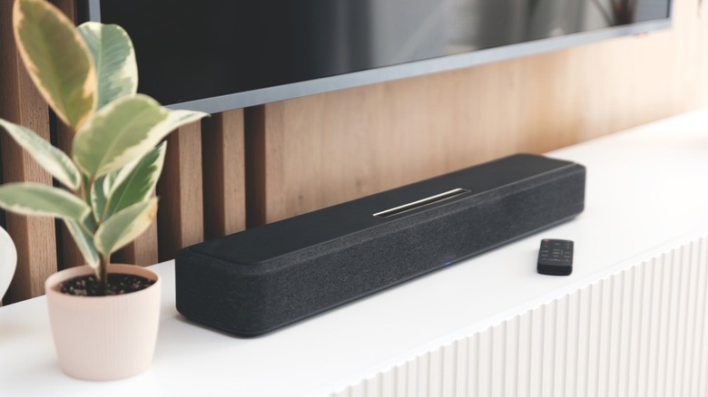 A soundbar sitting on a TV stand below a mounted TV with a plant next to it