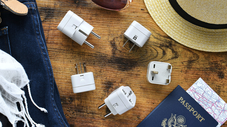 Several travel adapters on a wooden surface with a passport and other travel essentials.