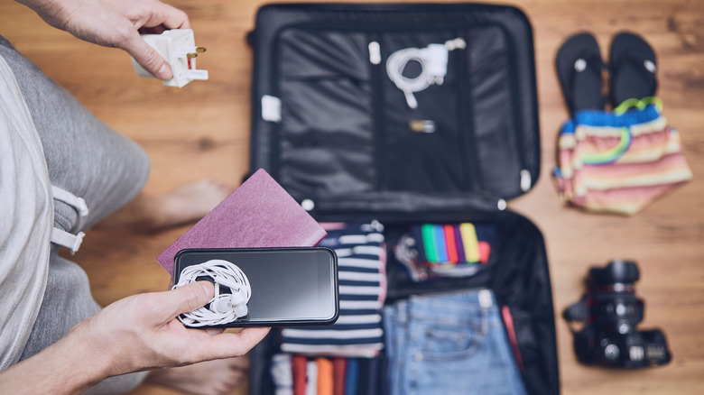 A traveler packing a suitcase with a smartphone, earphones, a travel adapter, a camera, and other travel essentials.