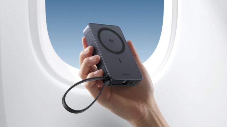 A hand holding a Ugreen MagFlow 10,000 mAh Qi2 wireless power bank wbeside an airplane window.