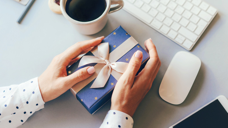 A pair of hands at a desk hold on to a gift in blue wrapping paper, a coffee cup, mouse, keyboard, and cellphone surround the gift