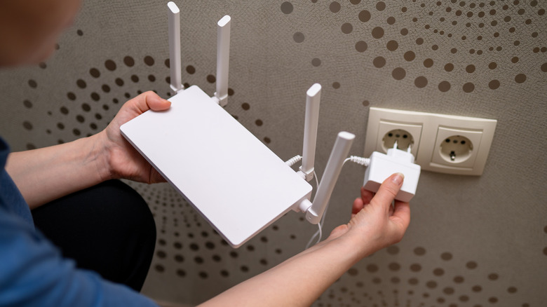 A person is connecting a wireless internet router to the electricity supply by plugging the power adapter into a wall socket.