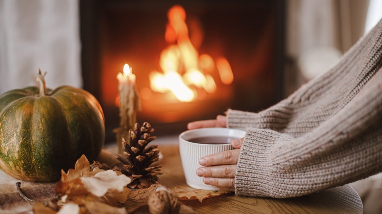 hands holding a cup full of liquid on an autumnal table with fire in the background