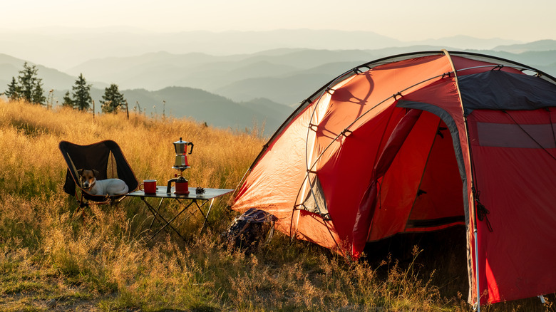 An orange tent sits on a hilltop next to a table and chair, there is a kettle on the table, as well as a large mountainous landscape in the background