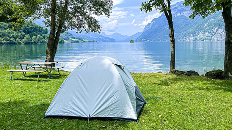 A silver tent sitting at a picturesque lakefront with a large mountain in the background, there is also a picnic table next to the tent