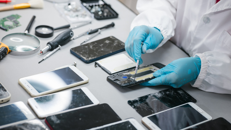 Several smartphones in various stages of repair sit on a table as a worker has a small screwdriver to the back of one of the devices, the worker is wearing a white lab coat and blue gloves.