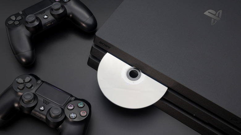 A black PlayStation 4 with a silver disc sticking out of its tray, two black Dual Sense controllers sit next to the system