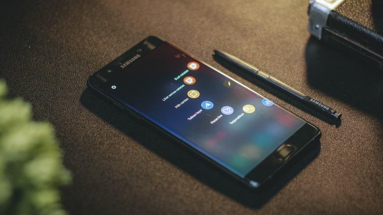 A close-up shot of a black Samsung Galaxy Note FE (Fan Edition) smartphone lying on a dark textured surface. The device's screen is active