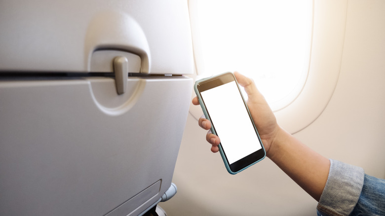 A hand holding a smartphone near an airplane seatback.
