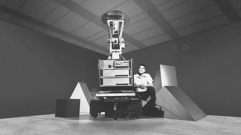 Black and white photo from the 1960s of a man crouching near a tall computer device, Shakey the Robot.