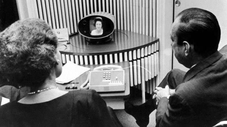 A black and white photo of two people looking at a screen with First Lady Lady Bird Johnson's face on it. They are making an early video call on a device called the Picturephone.