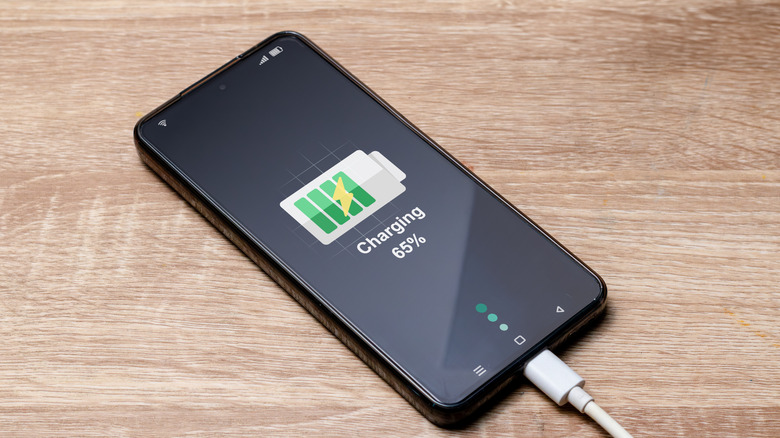 A phone connected to a charger showing the charging logo on its screen alongside text detailing it's 65% charged