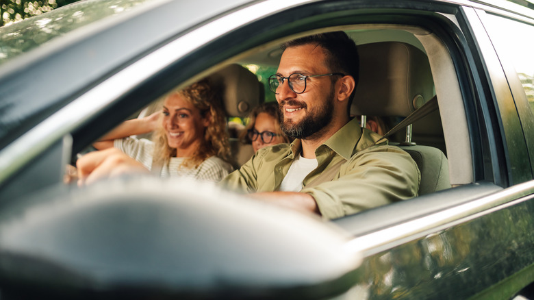 Person driving a car with passengers smiling in the back seats.