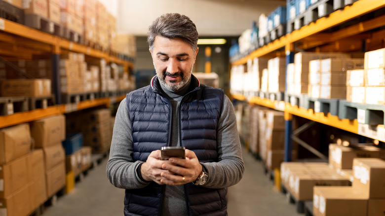 Man using a smartphone while standing in a warehouse aisle lined with boxes.