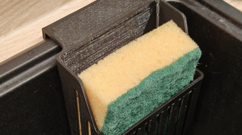 A green and yellow dish cleaning sponge being held in a Sponge drainer hooked onto a kitchen sink