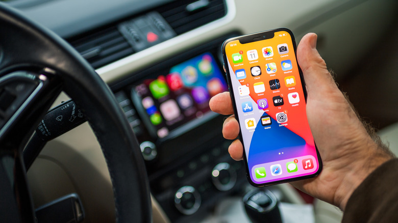 A driver holding an iPhone 12 Pro smartphone with a car infotainment system in the background.