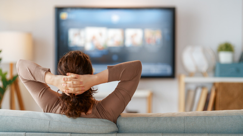 An individual with their hands behind their head sits on a light aqua couch while they watch TV, the photo is taken from behind the individual
