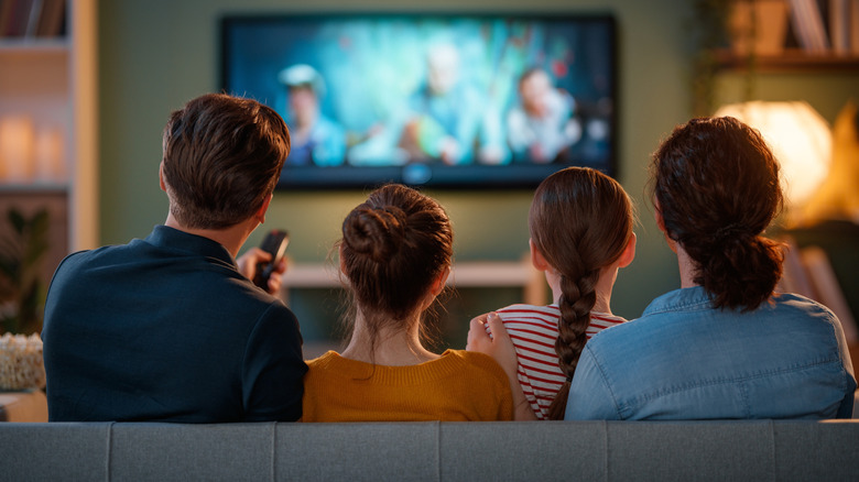 Two adults and two children are photographed from behind as they sit on a couch and watch TV in what appears to be a living room