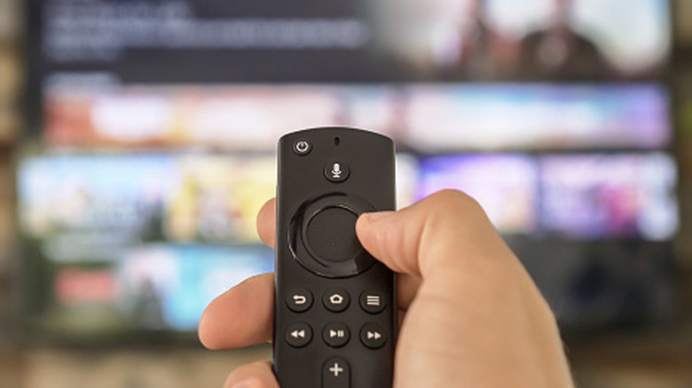 A closeup of a human hand holding a remote control as it's pointed at a TV, with the controller displaying the microphone option and control pad, among other buttons