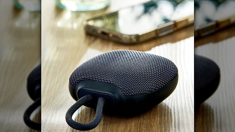 Rugged mini speaker placed on a wooden surface alongside a smartphone.