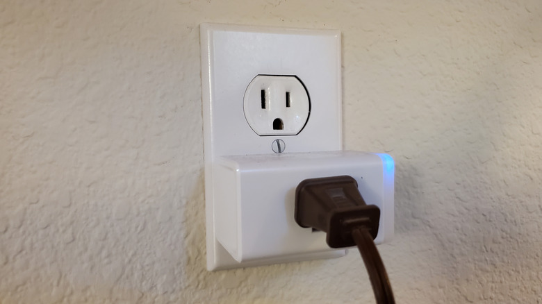 A black plug in a white smart plug that's in a white outlet on a white wall