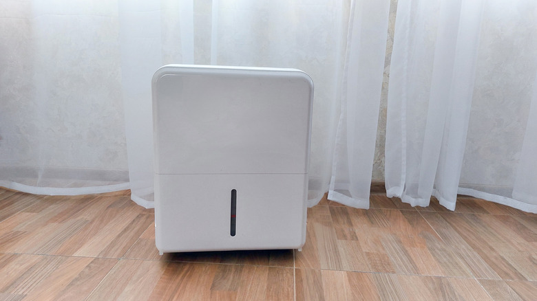 A white dehumidifier on a hardwood floor in front of white curtains