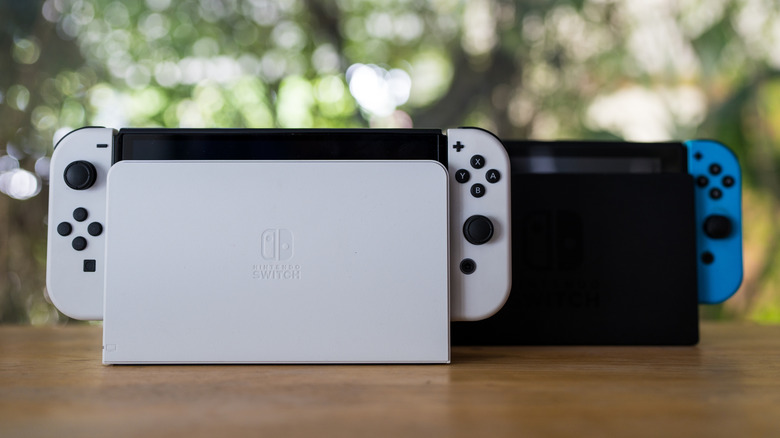 A Nintendo Switch with white Joy-Cons with black trim and a white dock sits next to a Nintendo Switch with a black dock and one blue Joy-Con visible