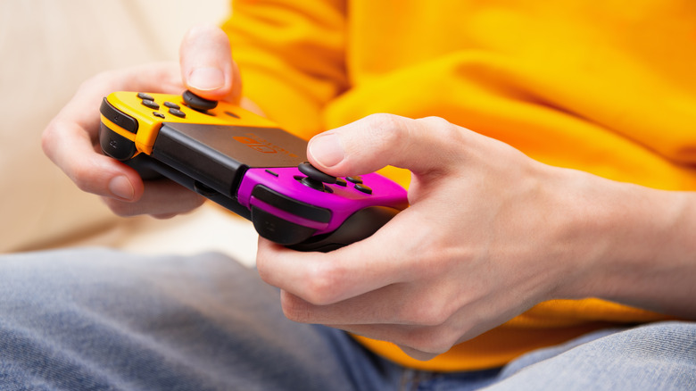 An individual in a bright yellow shirt with both of their hands on a Nintendo Switch Joy-Con controller, one Joy-Con is yellow and the other is a neon purple