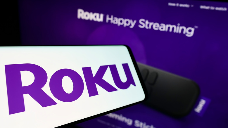 A cellphone prominently displaying the Roku logo is held in front of a screen displaying a Roku remote and the phrase "Roku Happy Streaming"
