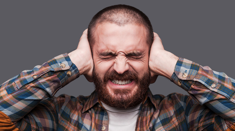 A bearded man in a plaid shirt squeezing his eyes shut and covering his ears with both hands against a plain gray background.