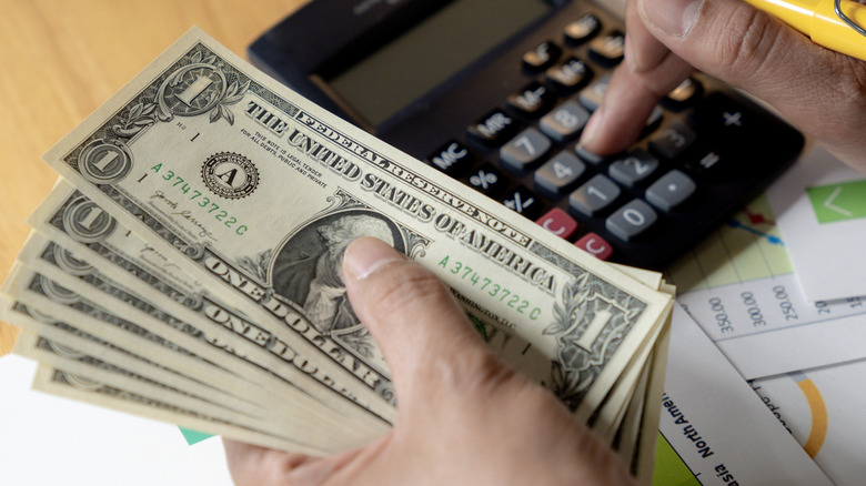 A close-up of a person counting U.S. dollar bills while using a calculator, with financial charts and paperwork visible underneath.