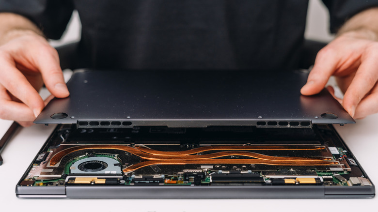 A person removing the bottom cover from a laptop, revealing internal components, including cooling fans and copper heat pipes.