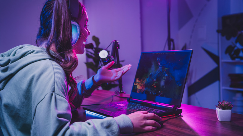 A person wearing a gaming headset with a microphone while playing a video game on a laptop in a room with purple and blue neon lighting.