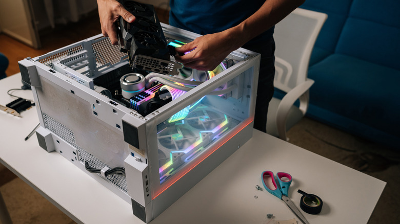 A person installing a graphics card into a white desktop PC case with RGB lighting, near tools and screws sit on the same desk.