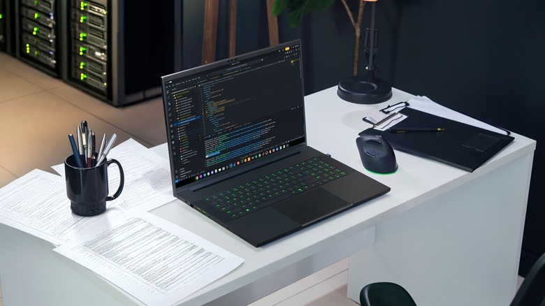 A diminutive modern laptop displaying code on a desk beside a wireless mouse, papers, and office supplies, with server racks visible in the background.
