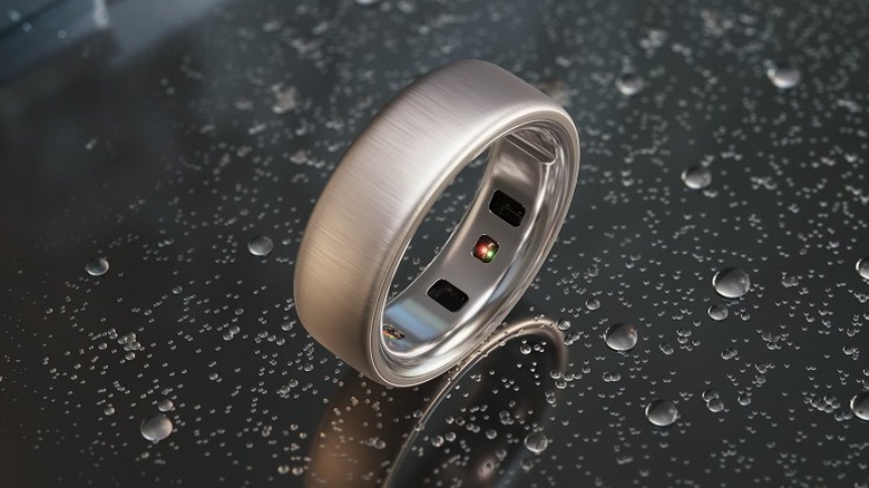A brushed silver Oura Ring 4 with its sensors visible placed on a dark surface with water droplets on it.