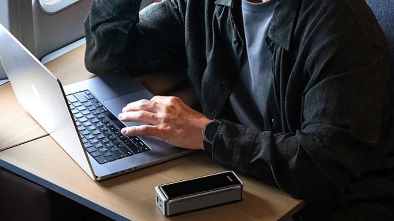 Man on an airplane working on a laptop with power bank and DSLR on the table.