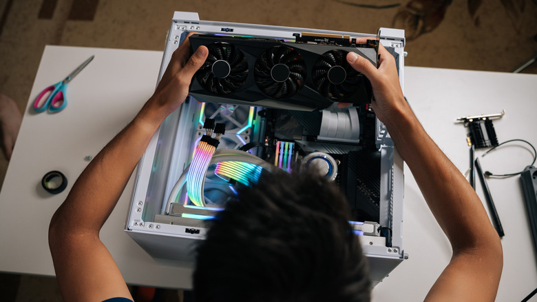 Top-down view of a person installing a large triple-fan graphics card into an open desktop PC with RGB-lit components and computer tools nearby.