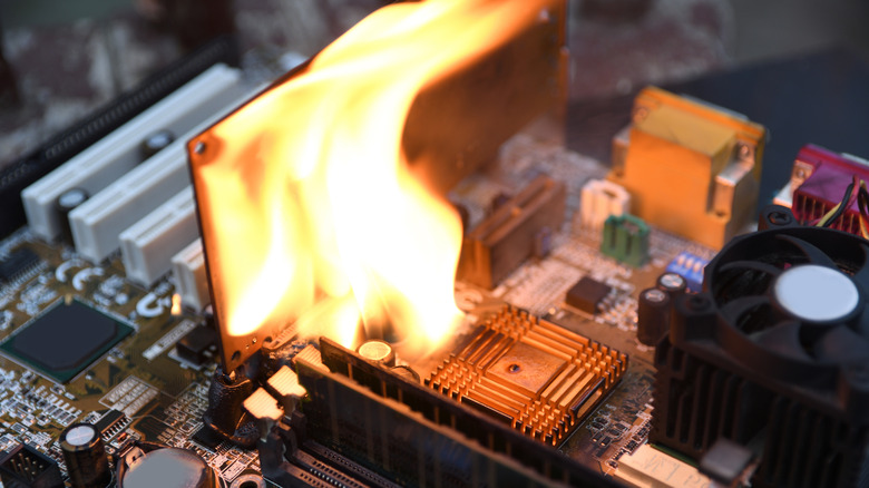 Close-up of a computer motherboard with flames rising from a component near the memory slots, while nearby heatsinks and a cooling fan remain visible.