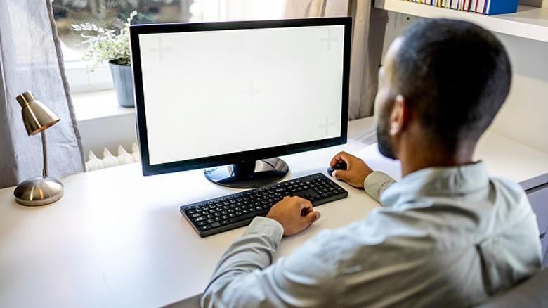Rear view of a person sitting at a desk and using a desktop computer with a completely white blank screen in a home office setting.