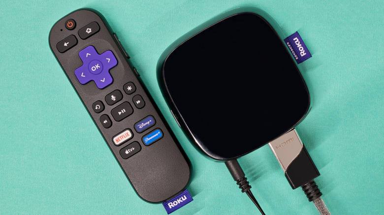 A Roku Streaming Device connected by its HDMI cable and power cable sits next to a black Roku remote against a sea foam green background