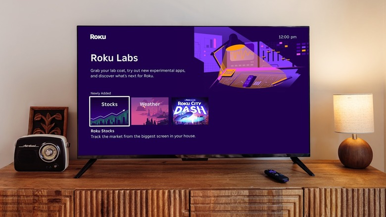 A television sitting on a wooden stand displays the new Roku Labs screen, offering users access to Stocks, Weather, and the "Roku City Dash" game