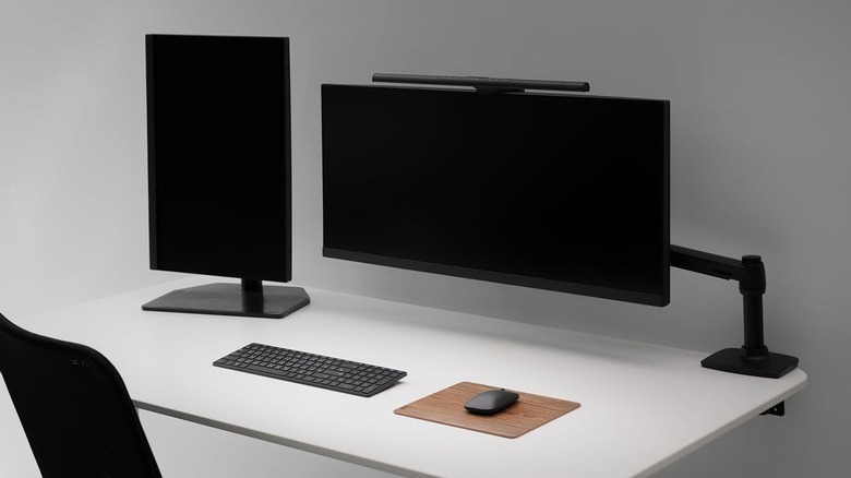 A minimal desktop setup with dual monitors, a curved ultrawide display on a monitor arm with a clip-on light, and a wireless keyboard and mouse on a white desk.