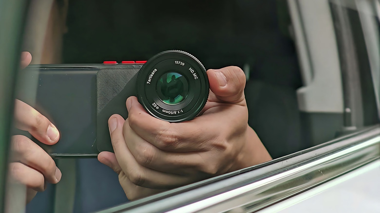 A person holding a smartphone with a large lens attachment, visible through an open car window.