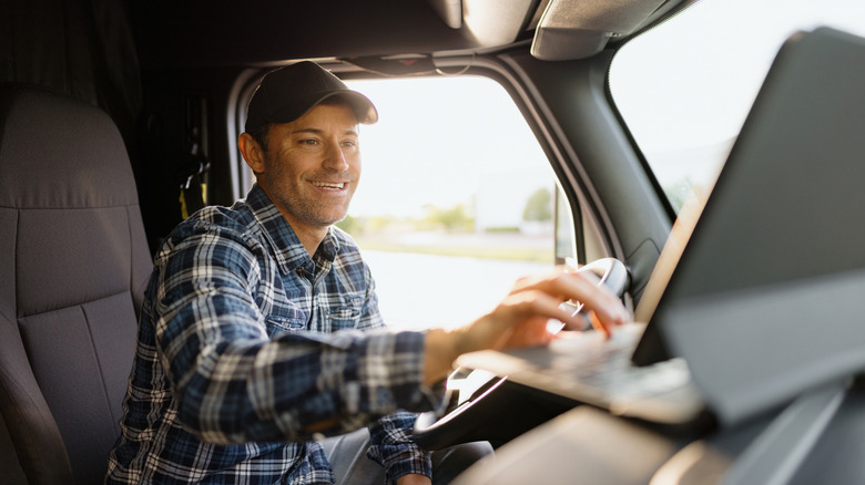 A truck driver smiling while reaching towards a laptop in his cab.