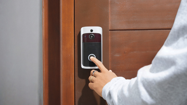 A person wearing a white sweatshirt reaching out to ring a doorbell camera on a wooden door.